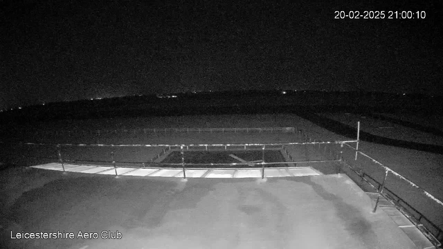 A dark scene from a webcam at the Leicestershire Aero Club, showing a flat rooftop with a railing. The area is dimly lit, and the faint outline of a runway is visible in the foreground. The sky is mostly black, with some distant lights hinting at surrounding structures or vehicles. The date and time displayed in the corner indicate it is nighttime.