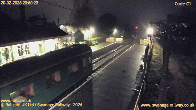 A dimly lit train station at night, featuring a green train on the left side of the image. The platform is wet, possibly from recent rain. To the right, there is a wooden fence and benches. The station building, partially obscured by shadows, has windows that glow warmly. A distant signal light can be seen in the background, casting a faint reddish hue. The overall atmosphere feels quiet and serene, with some tree branches illuminated softly by ambient light.