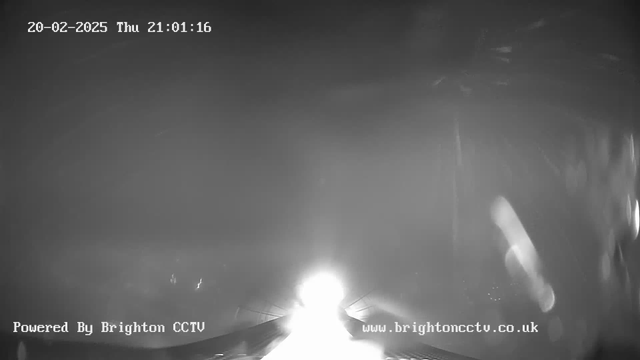 A black and white webcam image showing a bright light source in the center, creating a halo effect. The background is mostly dark with some lighter blurred areas. The top of the image displays the timestamp "20-02-2025 Thu 21:01:16" and the bottom includes the text "Powered By Brighton CCTV" and the website address "www.brightoncctv.co.uk". The overall scene appears to be nighttime.