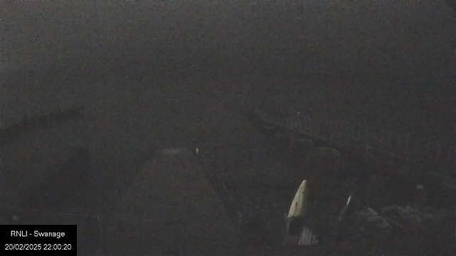 A low-light webcam image showing a dark scene. There are indistinct shapes in the foreground that appear to be structures or objects, possibly a railing or a dock. The background features a large, dark area that could represent water or an open space, blending into the night sky. There is minimal visibility across the scene, making it difficult to discern specific details. A timestamp indicating the date and time in the bottom left corner reads "20/02/2025 22:00:20."