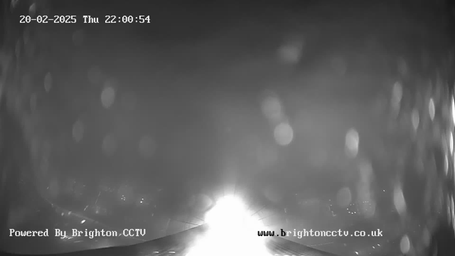 A black and white webcam image showing a bright light source at the center, surrounded by a blurred and hazy background. The image appears to have some drops or smudges affecting visibility, creating a distorted view. In the lower section, text reads "Powered By Brighton CCTV" and "www.brightoncctv.co.uk," indicating the source of the image. The date and time in the upper left corner show "20-02-2025 Thu 22:00:54."