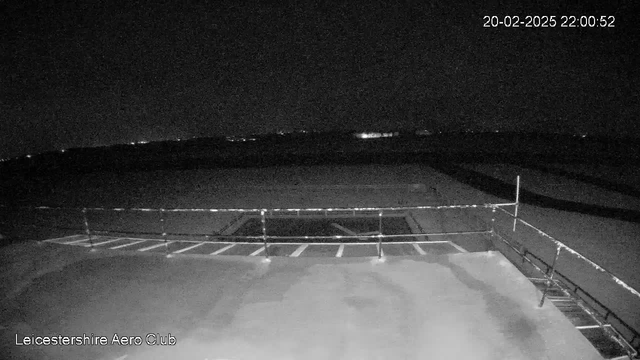 A dark scene captured by a webcam, showing an empty airfield at night. In the foreground, there's a flat surface with visible markings or lines, possibly indicating parking spaces. A railing runs along the edge. In the background, faint lights can be seen in the distance, suggesting the presence of structures or roads beyond the airfield. The top right corner displays the date and time: February 20, 2025, at 10:00 PM.