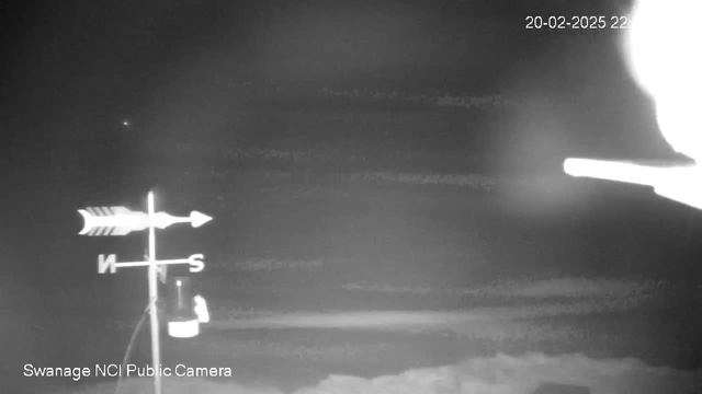 A black and white image of a night sky with low visibility, featuring a weather vane that includes directional arrows pointing North (N) and South (S). There is a faint light in the distance, possibly a star. The bottom left corner shows the text "Swanage NCI Public Camera." The date and time displayed in the top right corner is "20-02-2025 22:00." The overall scene appears dark and obscure.