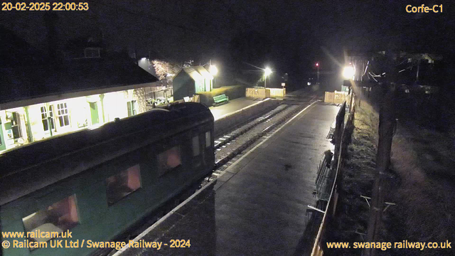 A dimly lit railway station at night, with a green train partially visible in the foreground. The platform features a bench and a small building with lighted windows. In the background, there are more benches and a waiting area, with a fence separating the platform from the surrounding area. Streetlights illuminate parts of the scene, but the overall atmosphere is dark.