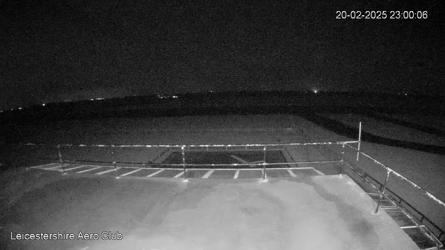 A dark scene captured by a webcam at night featuring a flat surface with a railing in the foreground. The background shows a large open area with a few distant lights visible, suggesting the presence of structures or vehicles. The image is monochrome, with limited visibility. The timestamp in the corner reads "20-02-2025 23:00:06."