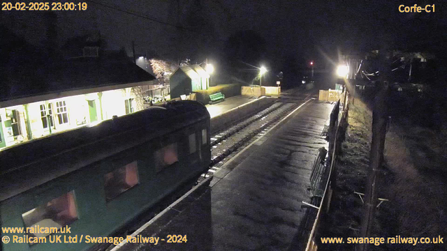 A dimly lit railway station at night. In the foreground, a vintage green train is partially visible, with its side showing several windows. The platform is empty and wet, reflecting some light. To the left, there is a building with large windows, illuminated from the inside. A bright light shines near the platform's edge, while a green bench is situated farther back. A wooden fence encloses part of the area, leading to a signpost and more light in the background.