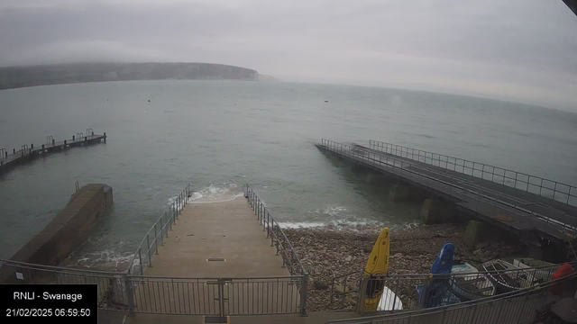 A foggy seaside view shows a concrete ramp leading to the water, with calm waves gently lapping against the shore. To the left, there is a small, rugged stone pier extending out into the sea, with several boat moorings. On the right, another pier is visible with a railing, leading to a flat surface. There are a few kayaks neatly stacked against the rocky shoreline, colored yellow and blue. The sky is overcast, creating a muted atmosphere, and the cliffs are faintly visible in the background.