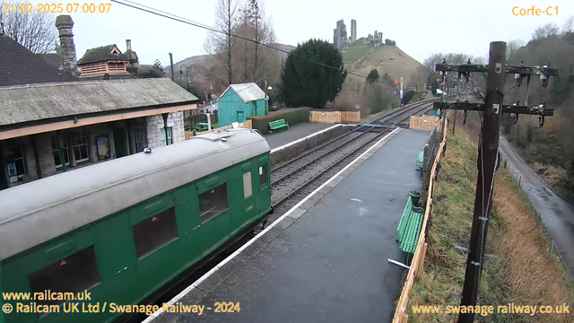 A green train car is parked at a station platform. The weather is overcast. In the background, there are distant hills with ruins of a castle. The platform features green benches and a wooden fence. An electric pole carrying wires stands to the right. A pathway leads away from the station.