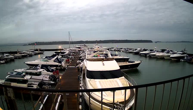 A marina filled with numerous boats docked at a wooden pier. The scene is set on a cloudy day, with a soft, muted light illuminating the water. Some boats are white and have various colored details, while others are darker. In the background, a distant shoreline is visible, surrounded by calm water. There are a few flags fluttering in the breeze, and the overall atmosphere is tranquil and somewhat overcast.