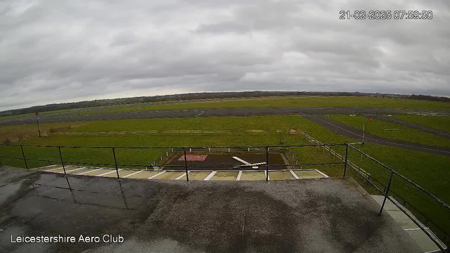 A cloudy sky spans the top of the image, with grey clouds dominating. Below, a grassy field extends across the scene, intersected by a black asphalt runway. A fenced area is visible in the foreground, housing a patch of red and white, possibly indicating a landing or takeoff zone. There are no visible aircraft in the image, and the overall mood is serene and empty, reflecting a tranquil morning at an airfield. The lower part of the image contains a ledge with a railing. The timestamp reads 07:59:50 on February 21, 2025.