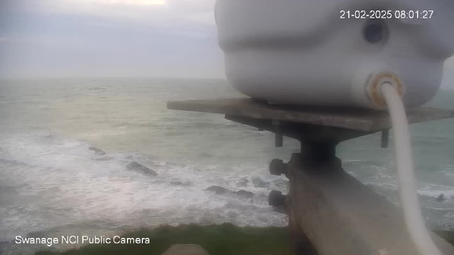 A cloudy sky is visible above a calm sea, with gentle waves lapping against the rocky shore. In the foreground, a white object is attached to a metal structure, possibly a camera or sensor, overlooking the water. The scene appears slightly hazy, suggesting a misty atmosphere.