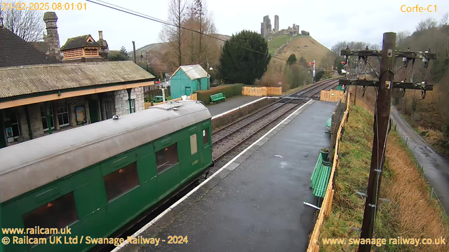 A view of a railway station in Corfe, showing a green train car approaching. The background features a hill with a castle ruins on top. In the foreground, there is a wet platform with a few green benches, a wooden fence, and trees. The sky is overcast with scattered clouds, and a power pole with electrical lines is visible on the right. The station building has a stone facade and a sloped roof.