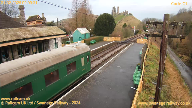 A view of a railway station featuring a green passenger train car parked on the tracks. The station building, made of stone with a sloped roof, is positioned to the left, with several windows visible. Behind it, a small turquoise building can be seen. In the background, a grassy hill rises, topped with a historical stone structure resembling ruins. The tracks extend into the distance, flanked by wooden benches in green and a wooden fence. A power pole stands to the right, with various wires overhead. The scene is cloudy and appears to be early morning.