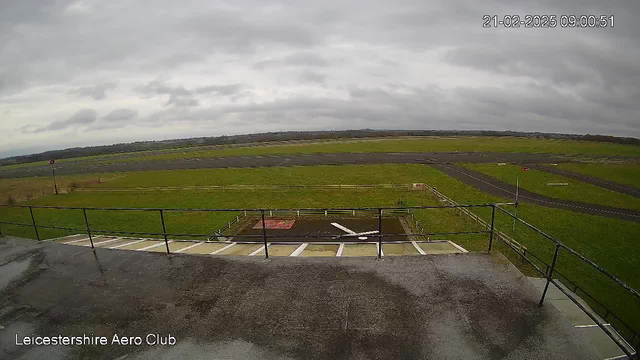 A view from a raised platform at the Leicestershire Aero Club, showcasing a wide expanse of green grass and a runway. The sky is overcast with gray clouds. In the foreground, there is a railing and a small area with a red marker on the ground. The runway extends in the background, bordered by a fence. A few landing lights are visible along the runway. The timestamp at the top right indicates the image was taken on February 21, 2025, at 9:00 AM.