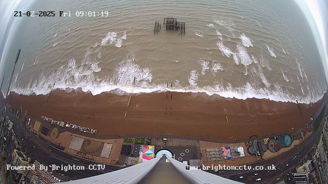 A view from above showing a wide sandy beach bordered by dark blue waves. The shoreline has several wooden piers, with one partially submerged in the water. In the lower portion of the image, there are colorful structures, likely beach attractions, along with a circular pattern and a pool. The sky is mostly cloudy, and the time stamp at the top indicates it was taken at 9:01 AM on February 21, 2025.