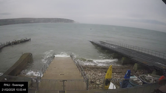 A view of a seaside location with a rocky shore and steps leading down to the water. The ocean is visible, with waves gently crashing against the shoreline. In the foreground, there are yellow and blue kayaks next to the steps. A dock extends into the water on the right, and a second dock is visible on the left. The sky is overcast, indicating a cloudy day.