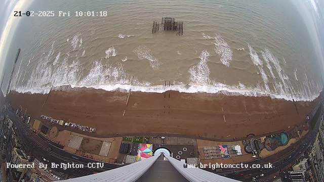 A wide view of a beach with waves gently crashing onto the shore. The scene is taken from above, showing a brown sandy beach with a partially submerged pier visible in the water. To the left, there are structures and attractions along the beachfront, including colorful sections and a circular design on the ground. The sky is overcast with light clouds, and the scene is timestamped with the date and time at the top of the image.