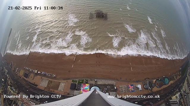 Aerial view of a beach with waves gently rolling onto the shore. The sandy beach is visible, with a small number of people walking along the water's edge. In the distance, an old pier structure is partially submerged. The area near the beach features colorful attractions, including tents and structures. A road runs parallel to the beach. The sky is overcast, with some light reflecting on the water. The timestamp shows it is February 21, 2025, at 11:00 AM.