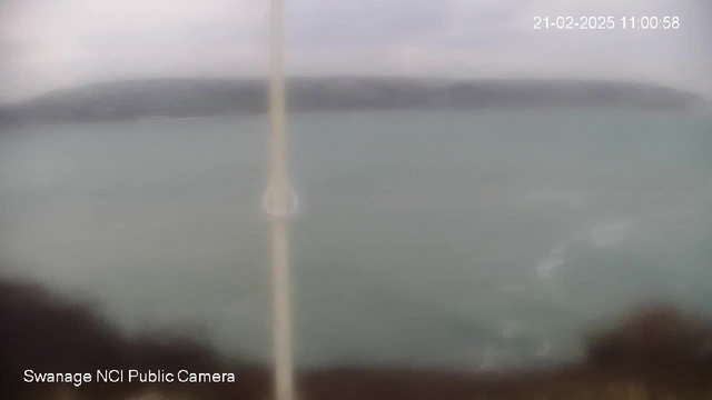 A blurred view of a seascape, with a pole on the left side. The water appears to be in shades of blue and green, and there is a distant coastline visible in the background under a cloudy sky. The date and time are displayed in the upper right corner.