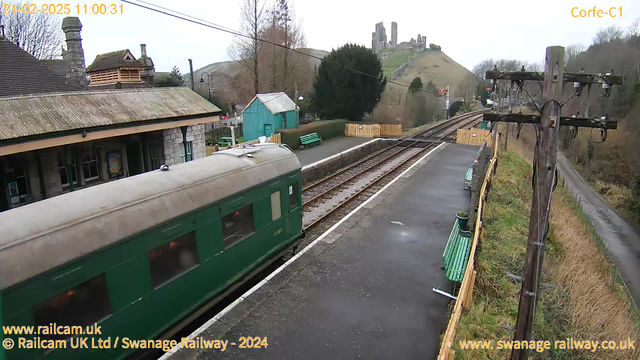 A green train car is parked at a quaint train station with a stone building, featuring a sloped roof and decorative wooden elements. In the background, a hill rises with the remains of a castle visible at the top. Scattered around the station are green benches and a blue wooden shed, while railway tracks extend toward the horizon. The scene is set on a cloudy day, with trees lining the station and hills in the distance. A power line pole stands to the right, adding to the rural charm of the setting.