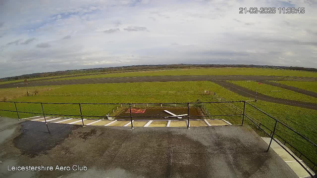 A view from a webcam at the Leicestershire Aero Club, showing a grassy airfield. In the foreground, there is a railing along a platform, and a few steps leading down. The airfield features several patches of grass, a runway marked with lines, and sparse trees in the background. The sky is mostly cloudy with some blue areas visible. Various ground markers and objects can be seen on the grass. The image has a timestamp indicating the date and time it was taken.