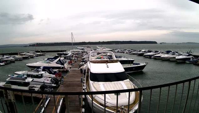 A view of a marina with numerous boats docked in the water. In the foreground, there is a large white motorboat with a striped top. Surrounding it are smaller boats of various sizes and colors, including blue and green. The marina is lined with wooden docks extending into the water. The sky is overcast with gray clouds, and a distant shoreline is visible in the background, partially obscured by the haze. The overall atmosphere appears calm and serene.