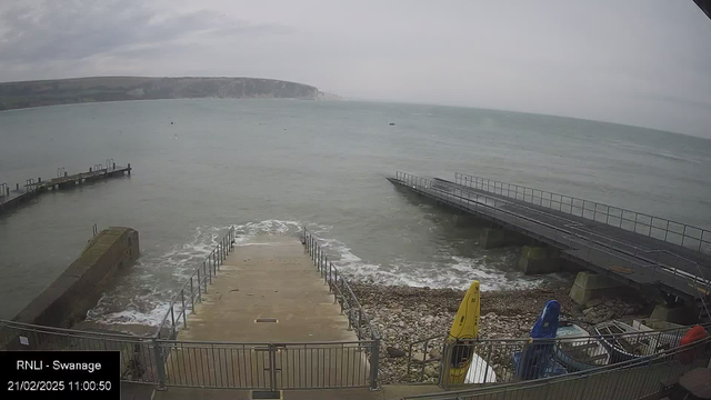 A view from a webcam showing the coastline at Swanage. In the foreground, there is a set of concrete steps leading down to the water, bordered by a metal railing. Waves gently wash onto the shore. To the left, a concrete jetty extends into the water, while a wider pier is visible on the right. Several colorful kayaks are lined up on the shore, with a yellow, blue, and red one prominently displayed. The sky is overcast with grey clouds, and the distant cliffs are partially visible in the background.