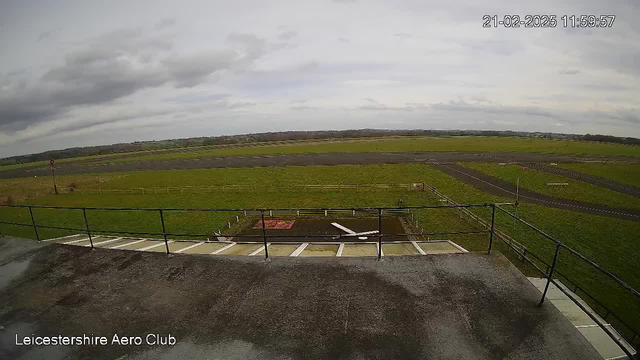 A view from a webcam at Leicestershire Aero Club shows an expansive green field and an airstrip. The sky is partly cloudy and overcast. In the foreground, there is a railing along a platform. Below, there is a small white object on the ground, likely a model aircraft. The airstrip is visible, running horizontally across the green landscape, surrounded by wooden fencing. The date and time are displayed in the corner of the image.