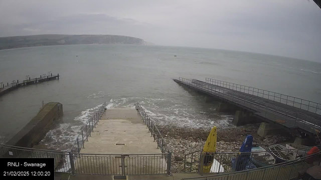 A view of a cloudy seascape showing a harbor area. Steps lead down towards the water, with waves lapping against the shoreline. On the left, a rocky structure extends into the sea. On the right, a wooden pier juts out over the water, and several brightly colored kayaks (yellow and blue) are positioned on the shore. The date and time are displayed in the bottom left corner.