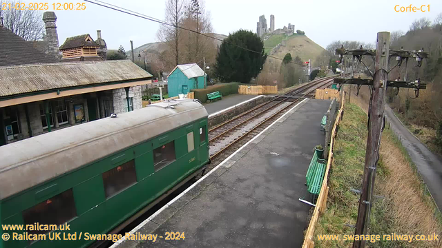 A green train car is partially visible at a train station. The station has stone buildings with a sloped roof and a chimney. There are green benches on the platform, and a wooden fence is visible in the background. In the distance, a hill rises with the ruins of a castle at the top. The sky is overcast, and there are trees lining the area. The image captures a rural setting, with train tracks running alongside the platform.