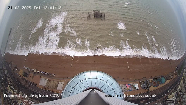 An aerial view of a beach with brown sand and rolling waves. The ocean is visible with white foam along the shoreline. A dilapidated pier structure stands partially submerged in the water. Below the camera, a circular glass observatory roof is visible, leading down to a paved walkway. There are various structures and vehicles along the beachfront, with some people along the sand. The sky appears overcast, adding a gray tone to the scene.