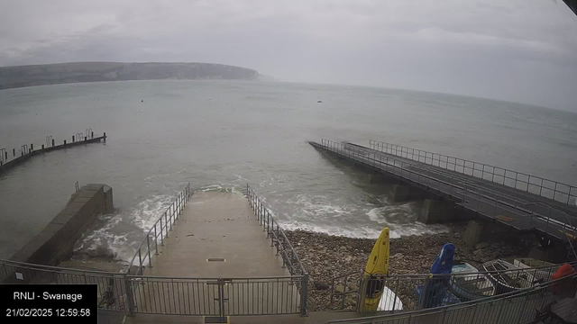 A cloudy view of a coastal area featuring a ramp leading down to the water. On the left, a small jetty extends into the sea, while on the right is a longer pier with a metal railing. The sea appears choppy with low waves, and there are a few boats nearby. Several kayaks, including a yellow one, are visible on the ground near the ramp. The background includes a steep, rocky shoreline and hills.