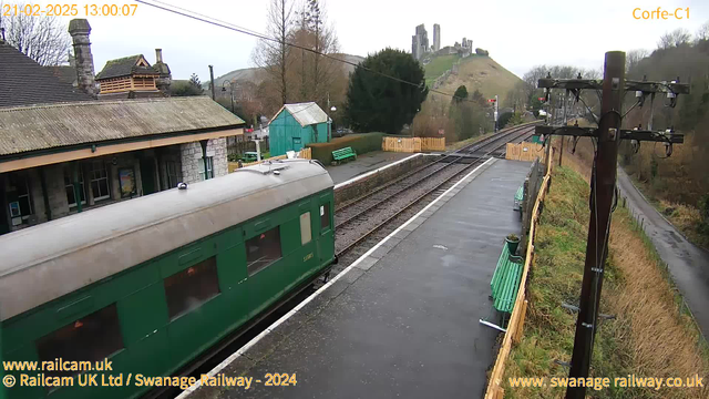 A green train carriage is prominently in the foreground, partially out of view. In the background, there is a railway station with a low stone building featuring a gabled roof. To the left, there is a turquoise building and several green benches along the platform. The tracks extend into the distance, passing by a wooden fence. On a hilltop to the right, there are ruins of a castle. The sky is overcast, giving a muted light to the scene.