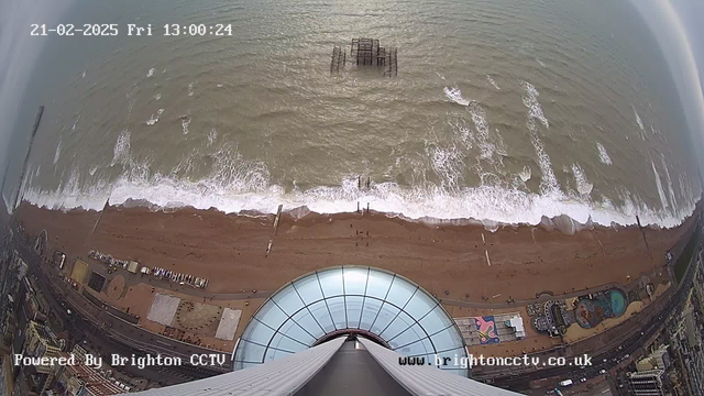 Aerial view of a coastline with a sandy beach and waves. The bottom of the image shows the edge of a circular glass structure. In the water, there are wooden posts visible, and further out, remnants of a pier can be seen. The beach is mostly clear, with a few structures and pathways. The sky is overcast, and the colors are muted, indicating a cloudy day. Time and date are displayed in the top left corner.