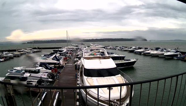 A marina scene captured from a webcam shows numerous boats docked in the water. Various types of boats, predominantly white yachts with some blue and other colored vessels, are lined up along a wooden dock. The water appears choppy, and the sky is overcast with gray clouds, creating a gloomy atmosphere. In the background, a coastline can be faintly seen, shrouded in mist. The image is slightly blurred, suggesting the presence of rain or moisture on the lens.