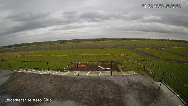 A view from the Leicestershire Aero Club webcam shows an open green field with a runway stretching across the center. The sky is overcast with clouds, creating a gloomy atmosphere. In the foreground, there is a railing along a terrace, and a small grassy area with a marked space resembling a landing pad. A fence lines the edge of the field, and there are a few other structures and markings visible in the distance. The scene conveys a quiet, expansive aviation setting.