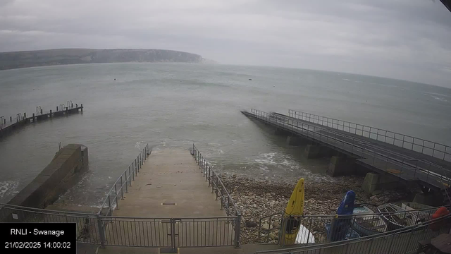 A view of a coastal area featuring a wide stretch of grey, cloudy sky above the sea, which appears choppy and greyish-blue. In the foreground, there are steps leading down to the water's edge, surrounded by a metal railing. To the left, a curved jetty extends into the water, while to the right, a flat, straight jetty is visible. There are two kayaks, one yellow and one blue, positioned on the shore. The area is rocky near the water, and the scene conveys a sense of calm despite the cloudy weather.