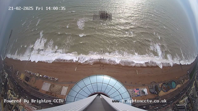 A high aerial view of the coastline shows sandy beach and rolling waves. The ocean appears choppy, with white foam at the crests of the waves. In the distance, there is a partially submerged pier structure. On the beach, there are several clusters of beachgoers and some beach equipment. Below the camera, the building features a large circular glass structure. The image is taken during the daytime, with overcast skies.