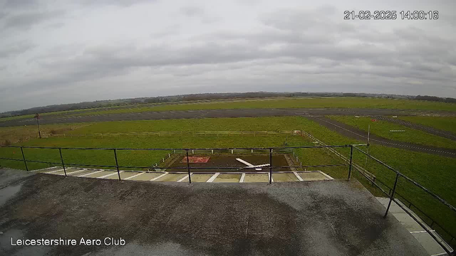 A wide-angle view of an airfield taken from a raised platform. The scene features a large expanse of green grass with a few patches of dirt. In the background, there is a gray, overcast sky. A runway, marked with dark asphalt and white lines, stretches across the field. In front of the platform, a circular area is partially visible, with a white object that resembles a plane component lying in it. A fence surrounds an area in the distance, and a small red structure can be seen on the edge of the airfield. The timestamp in the corner indicates the date and time of the image.