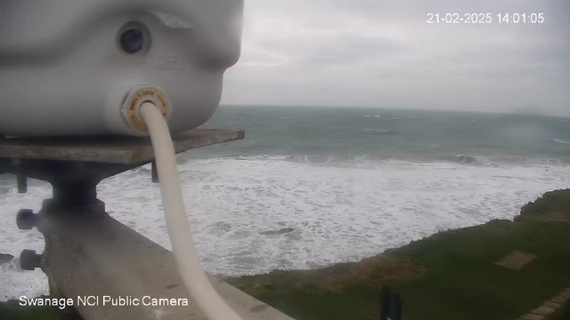 A coastal view showing choppy waves crashing against the shore, with a cloudy sky above. The foreground features a white device or camera mounted on a ledge, connected to a cable. The grass and gravel areas of the shore are visible in the lower part of the image.