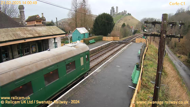 A green train carriage is positioned on the left side of the image, with a view of a railway station platform. The platform features benches painted in green and a stone building with a sloped roof in the background. In the distance, a hill can be seen topped with ancient ruins or castle remains. The sky is overcast, and the scene conveys a quiet, rural railway setting.