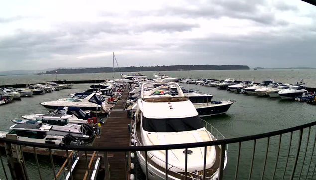 A view of a marina on a cloudy day, featuring numerous boats docked in the water. The foreground shows a wooden pier with several boats, including a large white yacht. In the background, more boats are visible on the water, surrounded by a gray sky with low clouds. The setting is peaceful, with gentle waves and distant land visible across the water.