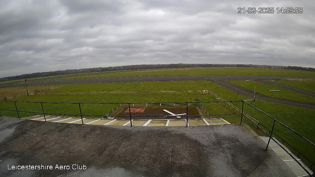 A wide view of an airfield under a cloudy sky. In the foreground, there is a railing indicating an elevated viewpoint. The ground is mostly green grass with a small section of bare earth, where a white propeller is resting. In the background, there are multiple runways, some marked with white lines. A red windsock stands to the left, and there are several wooden fences surrounding parts of the field. The scene appears tranquil and open, typical of an aeroclub setting.