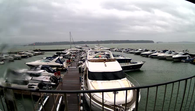 A marina scene with numerous boats docked in a harbor. The foreground features several motorboats and yachts, including a large white yacht with a blue canopy. The background shows more boats and a cloudy sky over the water, with a hint of land on the horizon. The overall atmosphere appears overcast and calm, suggesting a typical day at the marina.