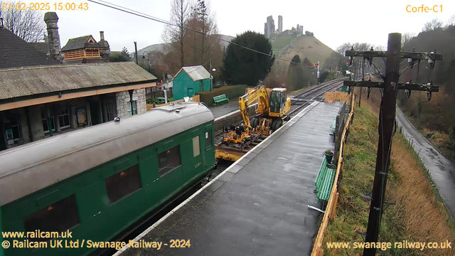 A view of Corfe Castle station on a cloudy day. A green train car is positioned on the left, with a yellow construction vehicle near the railway track, working on maintenance. The background features a row of benches and buildings, and the ruins of Corfe Castle are visible on a hill in the distance. The platform surface appears wet, indicating recent rain.