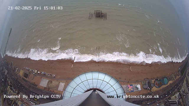 A high-angle view of a beach and the sea. The foreground shows a round, glass dome structure, possibly part of a building. Below the dome, the sandy beach stretches out, lined with various structures and vehicles. In the distance, waves are washing onto the shore, and a partially submerged pier is visible in the water. The sky is cloudy, and the image is timestamped with the date and time in the top left corner.