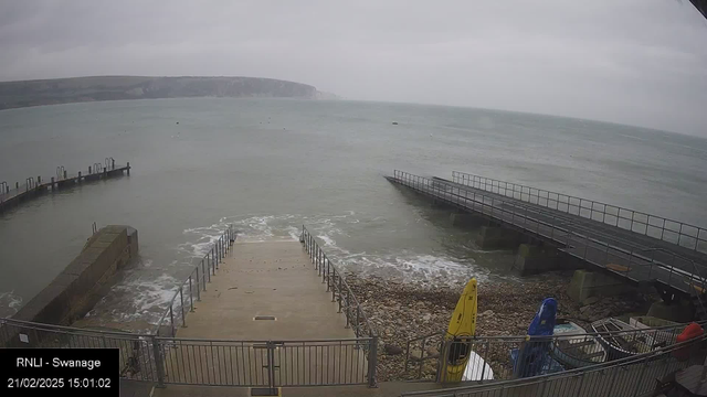 A coastal scene showing a grey sky above a choppy sea. Waves crash against a rocky shoreline, where a set of concrete steps leads down to the water. There are two wooden piers extending into the sea from the left side, with several boats visible on the shore, including a yellow kayak. The overall atmosphere appears calm but overcast, typical of a potentially chilly day by the sea.