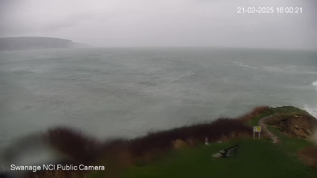 A cloudy, overcast seascape with turbulent waves crashing against a shoreline. The horizon features a distant cliff, while the foreground shows a grassy area with a bench and signage. The atmosphere appears stormy, with limited visibility.