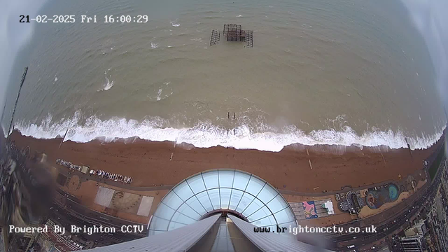 A high-altitude view overlooking a beach with a wide expanse of brown sand and a choppy blue-green sea. The waves are crashing against the shore, creating white foam. In the distance, remnants of an old pier structure can be seen partially submerged in the water. At the bottom of the image, there are colorful attractions and facilities along the beach promenade, including a circular pattern that appears to be a carousel or similar ride. The scene captures a cloudy atmosphere, indicating an overcast sky. The date and time are displayed in the top corner, with the text "Powered By Brighton CCTV" at the bottom.