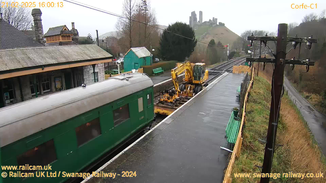 A railway station scene featuring a green train car on a wet platform. In the background, there is a yellow construction vehicle near the tracks. To the left, a station building with a stone exterior can be seen along with a few trees. On the hillside behind, ruins of a castle are visible. The sky is overcast. The platform has wooden benches and some greenery, while power lines run along the edge of the view.